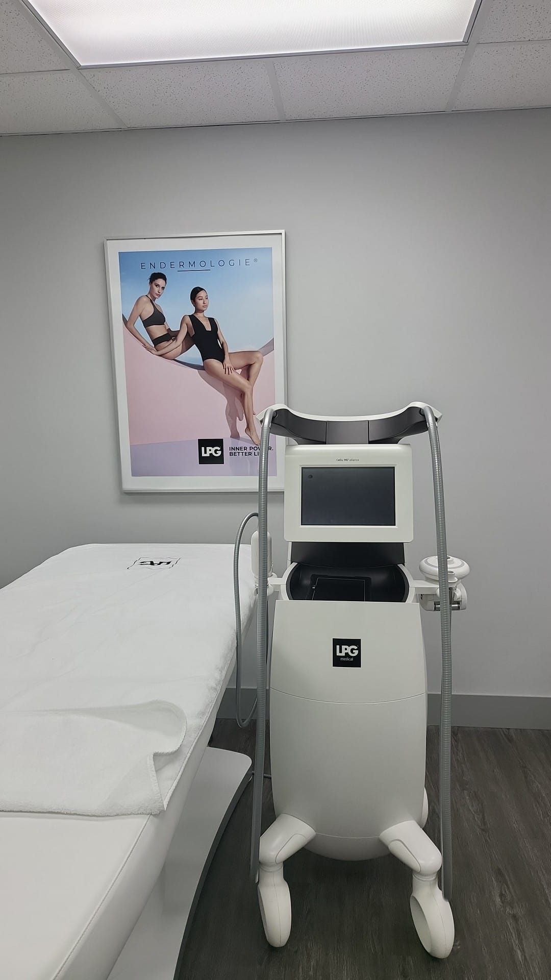 A medical examination room with a white bed, body contouring machine, and a poster featuring two women.