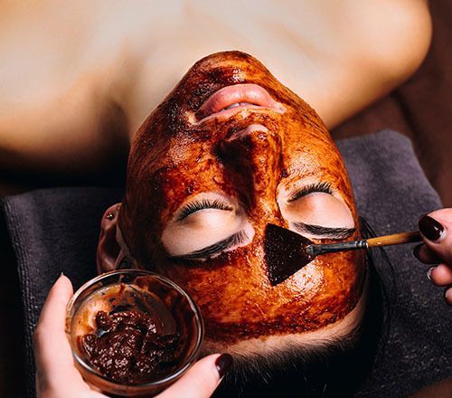 Woman receiving a facial with a brown mask. A hand applies the mask with a brush, holding a small bowl of mask mixture.