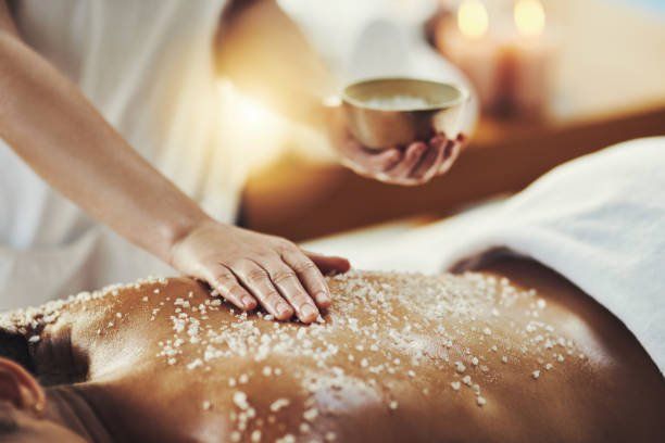 A person receiving a salt scrub on their back at a spa. A hand pours salt onto the person's back with lit candles in the background.