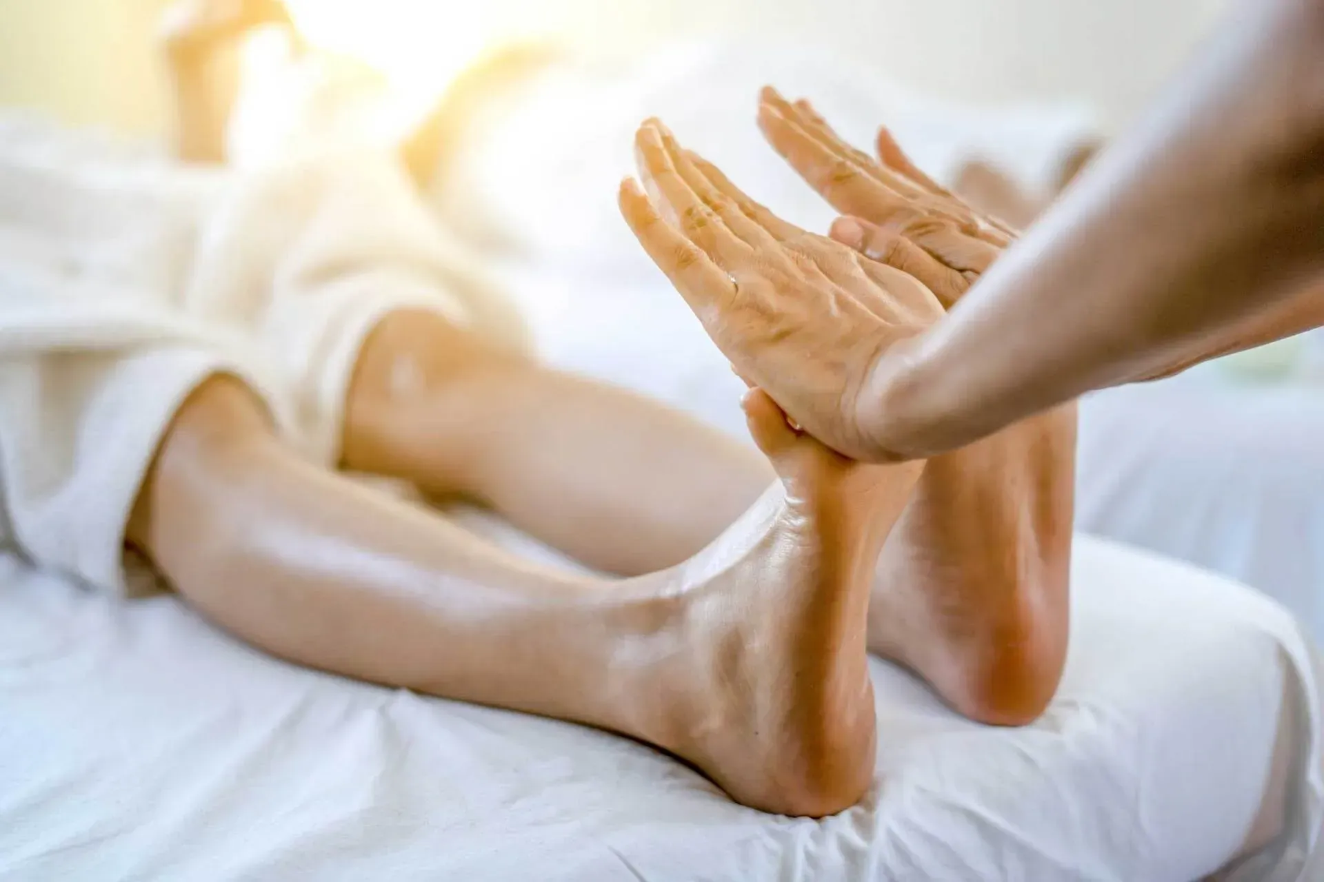 Hands massaging a person's foot on a white bed in a bright setting. The focus is on the foot and hands.