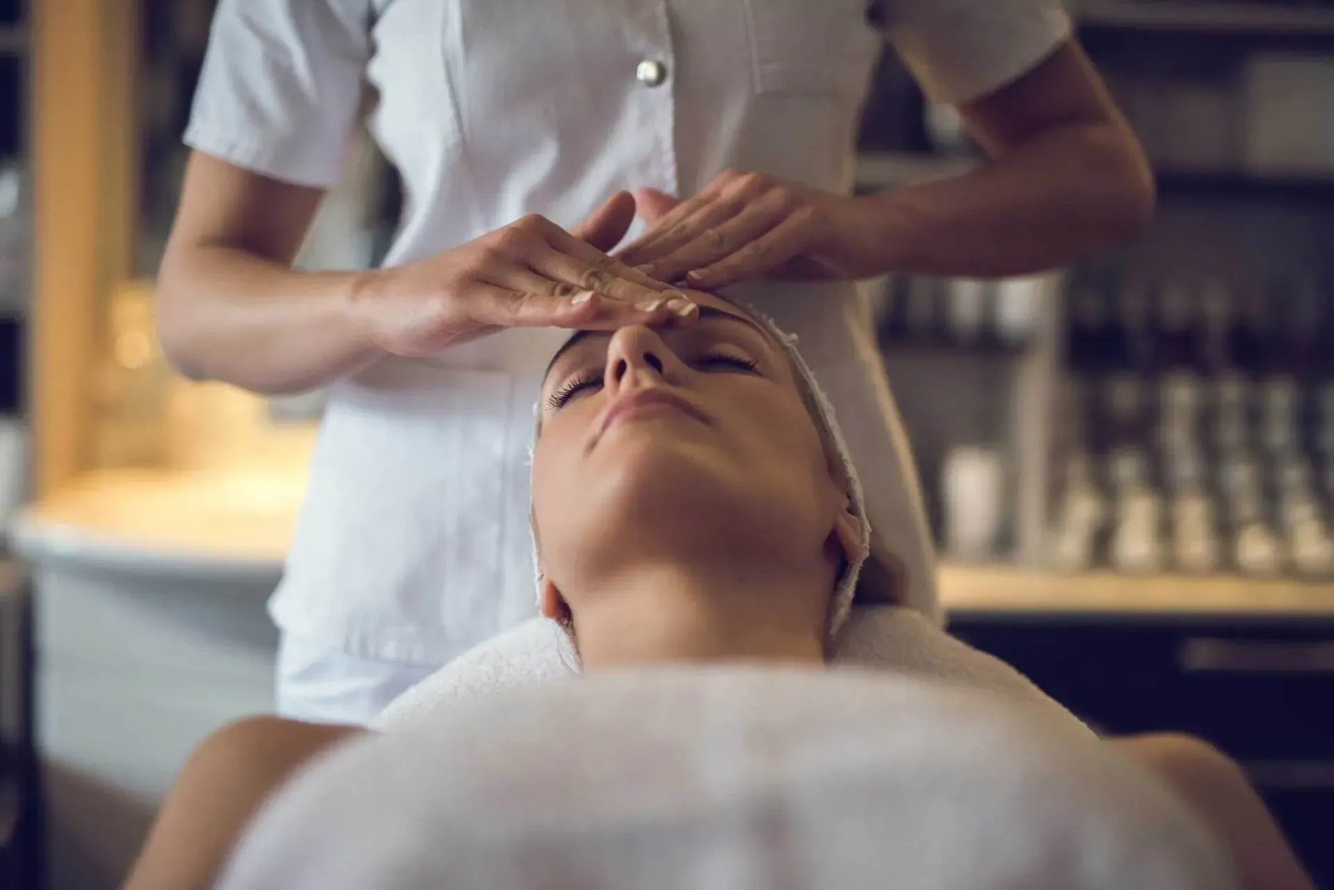 Woman receiving facial massage at a spa, lying on a towel with eyes closed. Therapist's hands are on the woman's forehead.