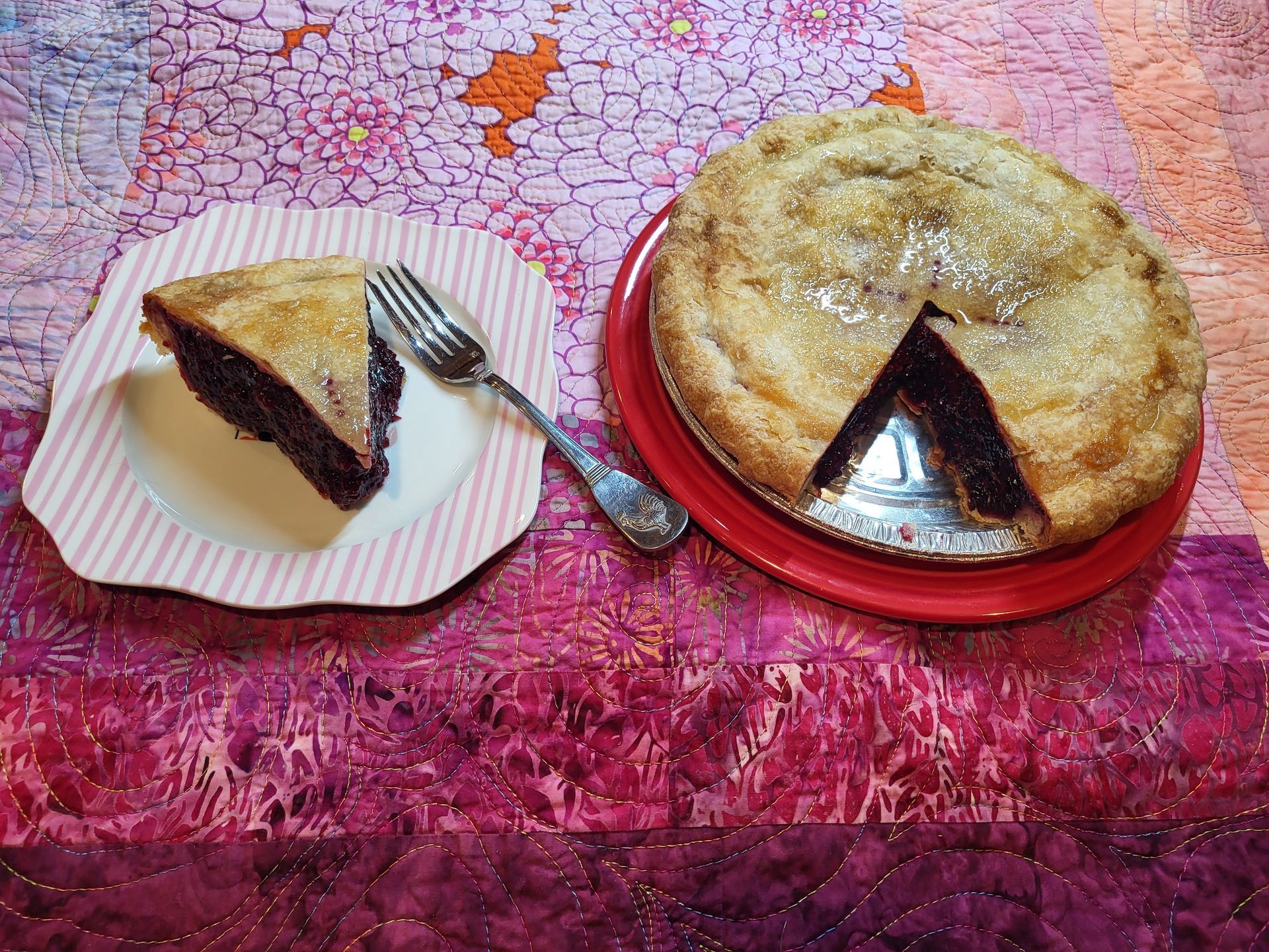 A berry pie with one slice served on a pink-striped plate with a fork, set on a colorful, patterned fabric surface.