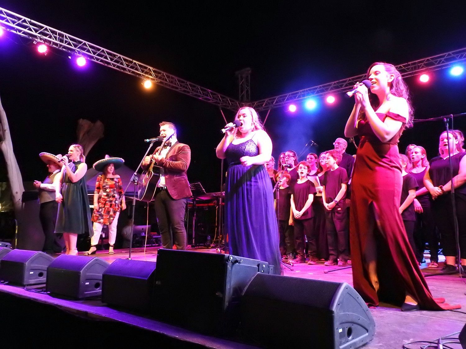 People singing on stage, under stage lights. Musicians and chorus in the background. Night setting.