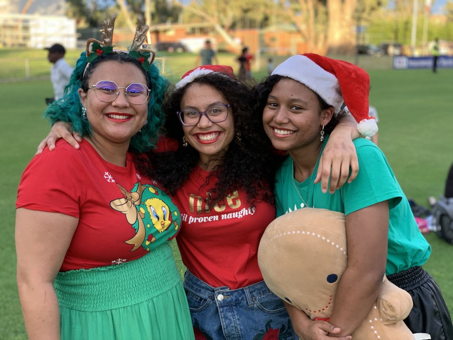 Three smiling women in Christmas attire pose outdoors on grass.