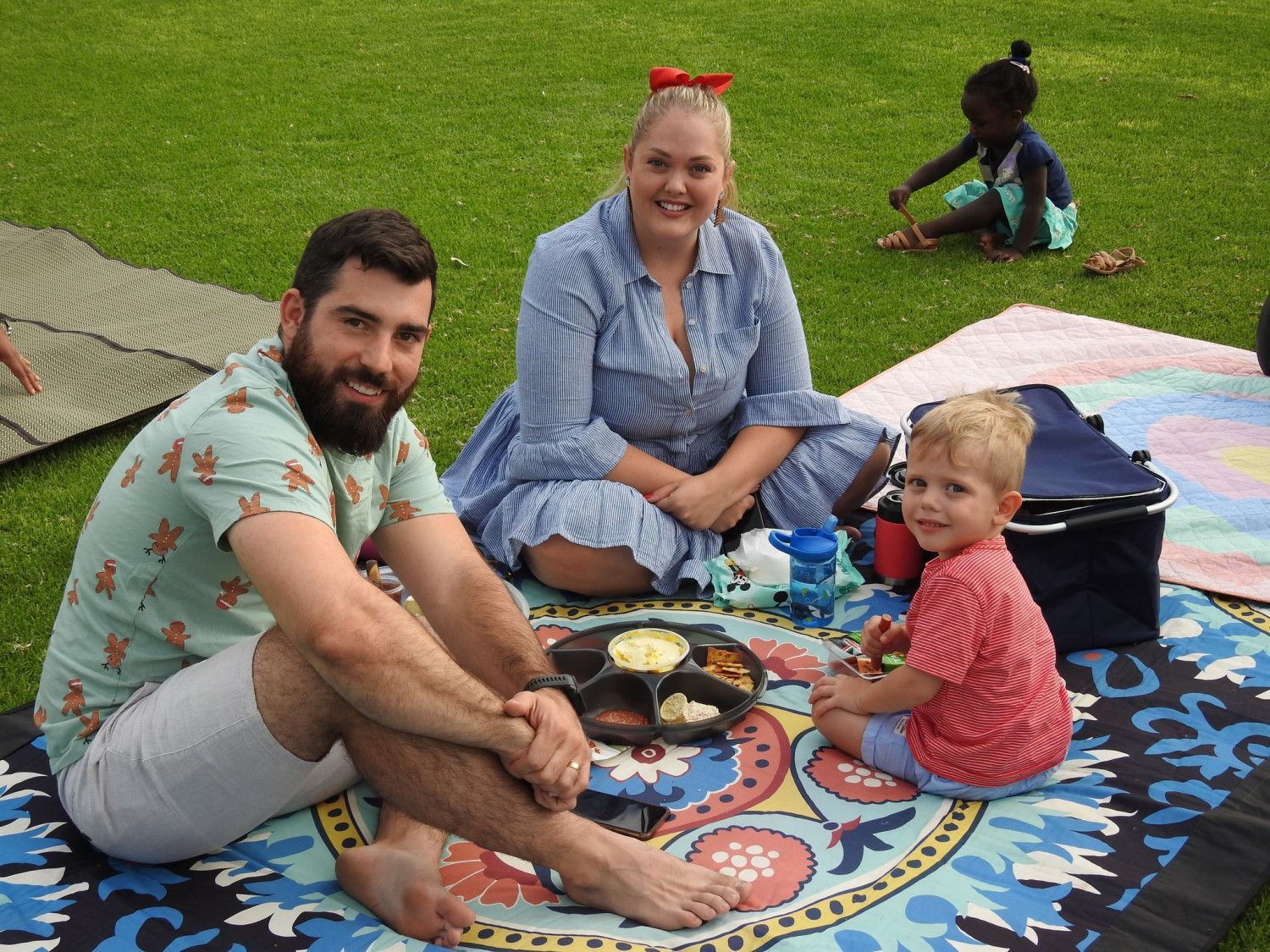 Family picnicking on a patterned blanket; man, woman, boy smiling, food on a plate. Child in the background.