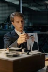 A person in a suit sits in a dim office, holding up a photograph of someone wearing glasses.