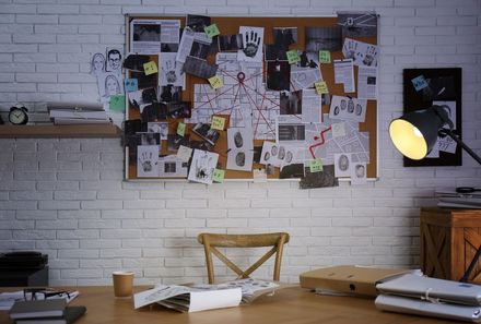 An investigator's workspace featuring a corkboard covered in documents, maps, and crime scene photos above a wooden desk.