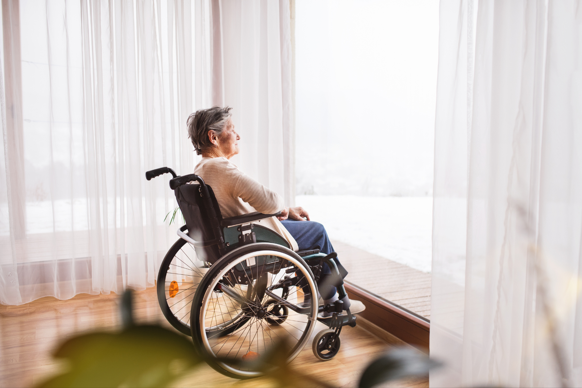 An elderly woman in a wheelchair is looking out of a window.