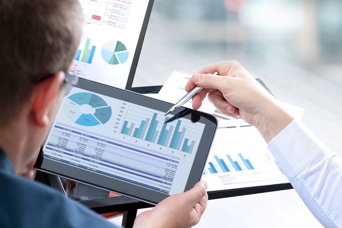 Una persona tiene in mano un tablet con grafici aziendali mentre indica un monitor desktop che visualizza grafici analitici.