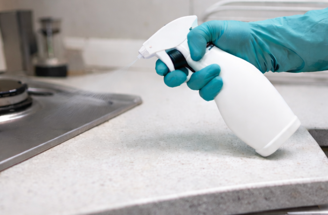 Gloved hand spraying cleaner on a kitchen countertop near a stovetop.