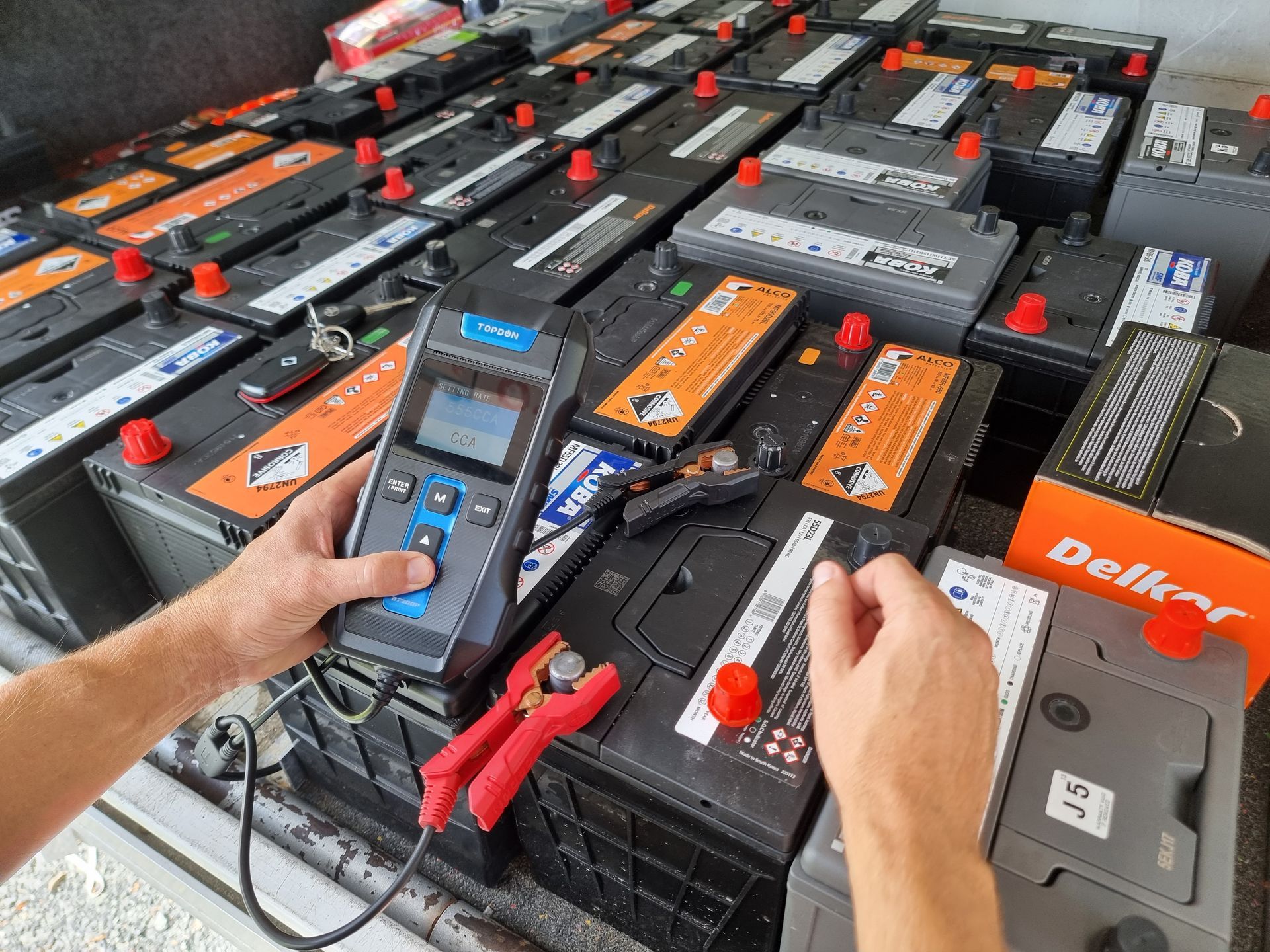 Hands testing car battery terminals with a digital device among many batteries. — ASAP Batteries & Breakdowns in Burleigh Heads, QLD