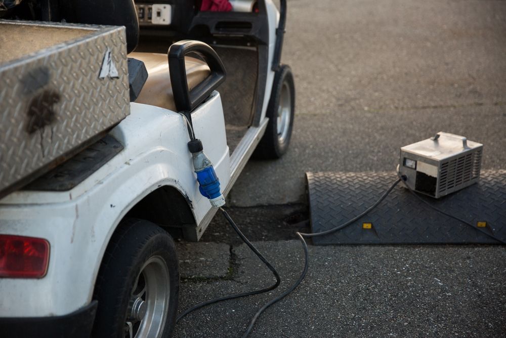 White golf cart charging with a cord connected to a metal box on a black mat on pavement. — ASAP Batteries & Breakdowns in Burleigh Heads, QLD