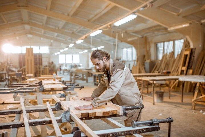 Carpenter working on wood in a workshop with tools, tables, and lumber.