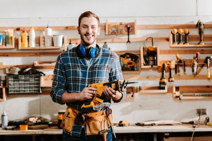 Smiling man in workshop holding saw, wearing ear protection and tool belt.