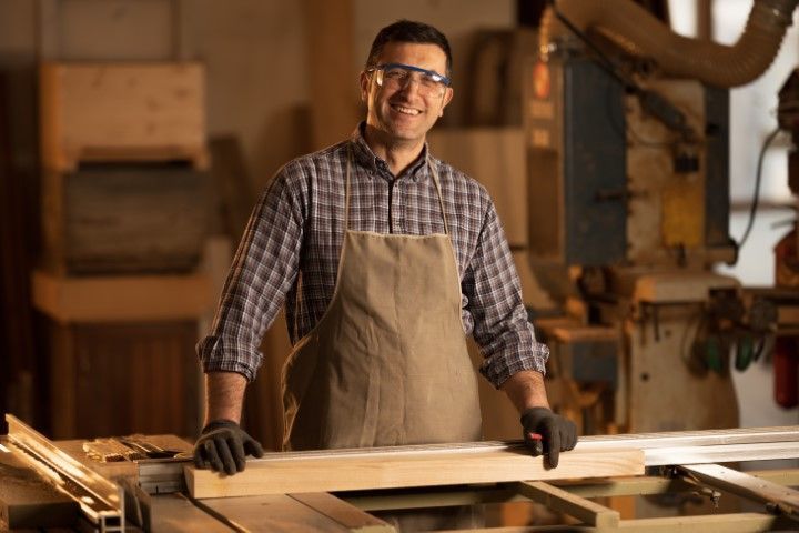 Smiling carpenter in apron and safety glasses, holding wood at a workbench in a workshop.
