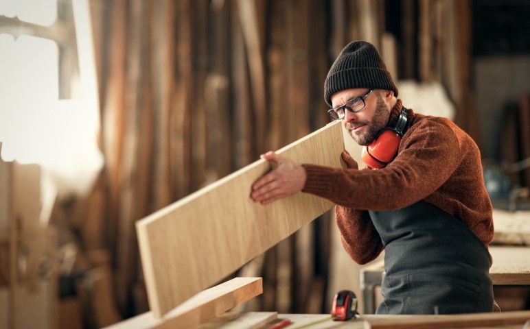 Carpenter in a workshop holding a wooden plank, wearing earmuffs and beanie.