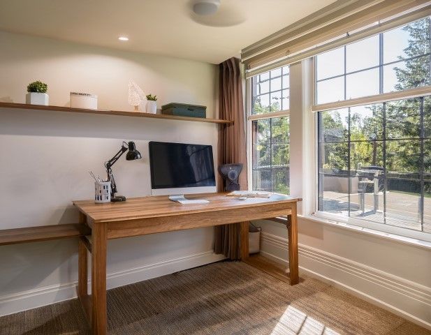 Wooden desk in front of a window, with a computer and plants. Sunlight fills the room.