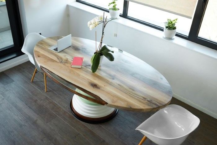 Oval wooden table with a laptop, plant, and books, flanked by white chairs in a bright room.