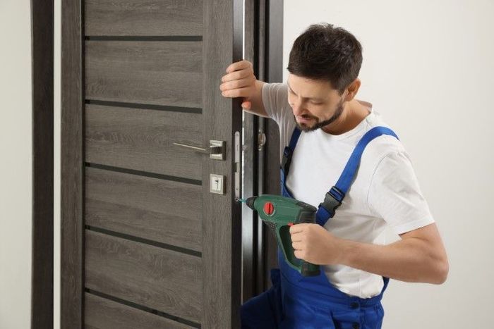 Person in blue overalls using a power drill to work on a gray door.