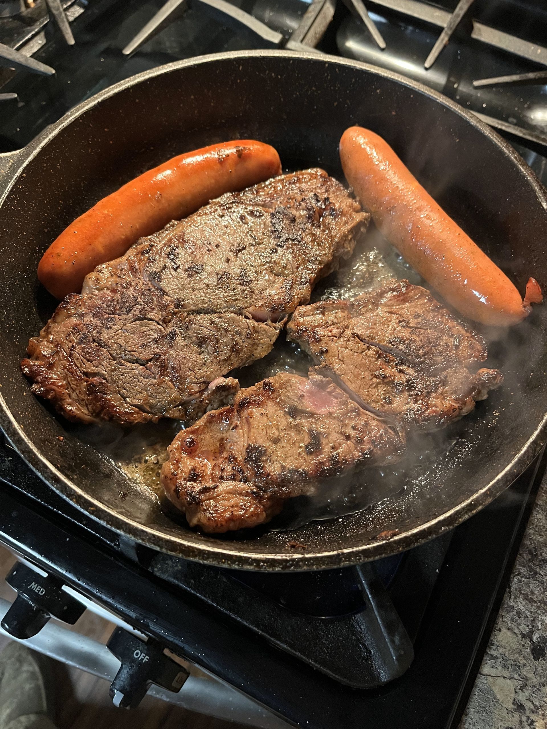 Steaks and sausages are cooking in a pan on a stove