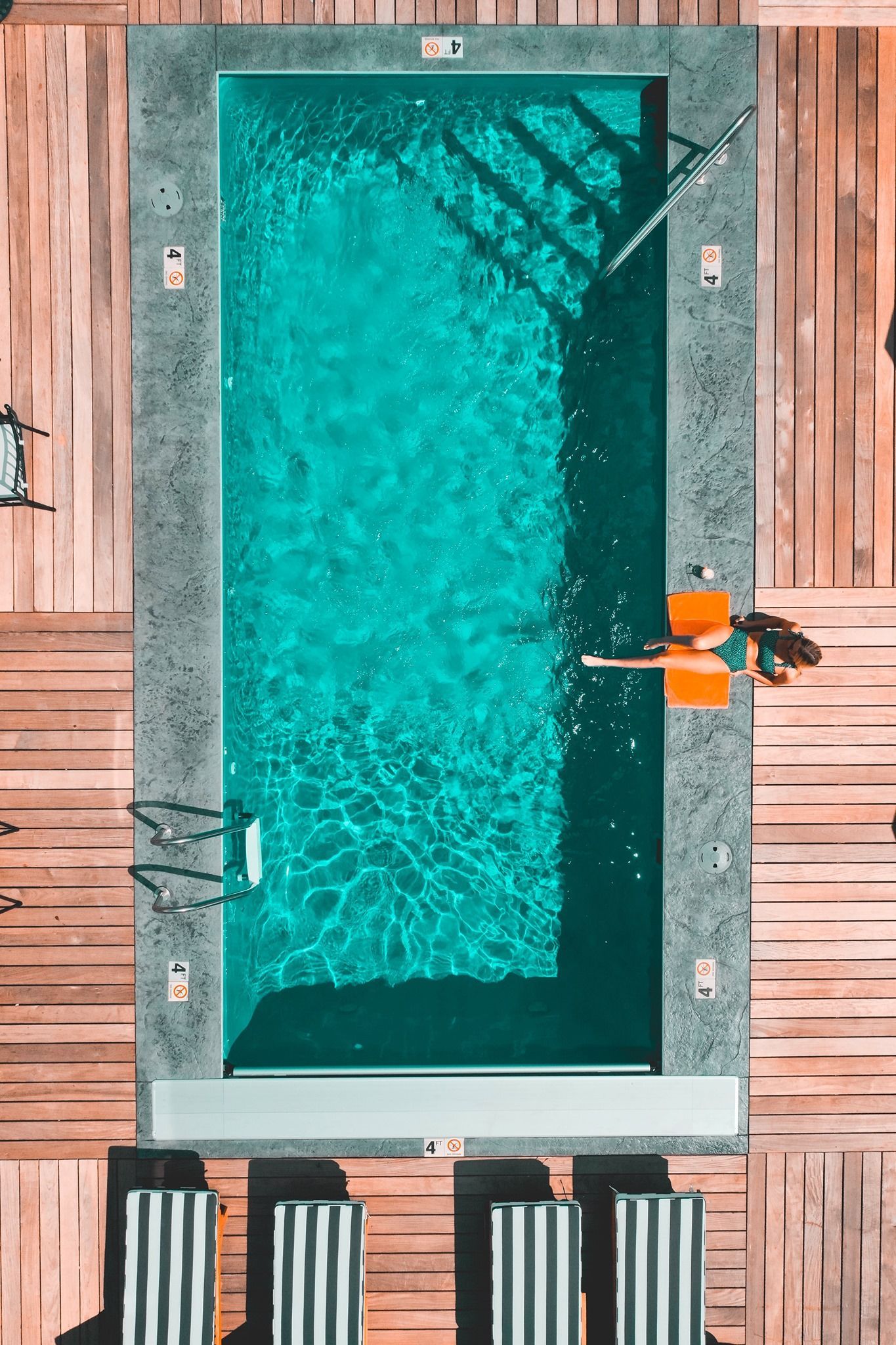 Aerial view: Rectangular turquoise pool with person on float, surrounded by wood deck and lounge chairs.