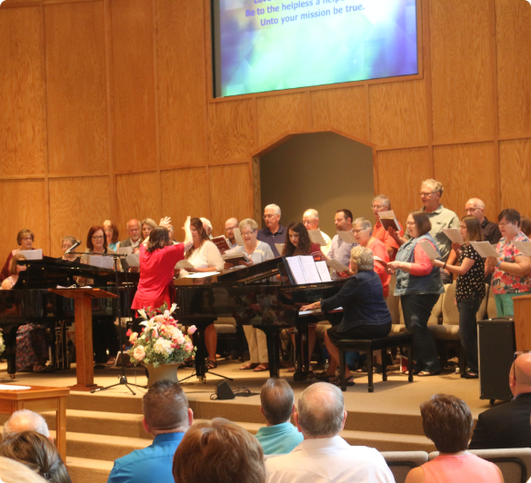 A choir stands on a stage with a grand piano and podium before an audience in a wooden-paneled church sanctuary.