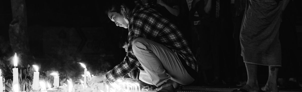 A person in a plaid shirt kneels in the dark, lighting a row of candles on the ground.
