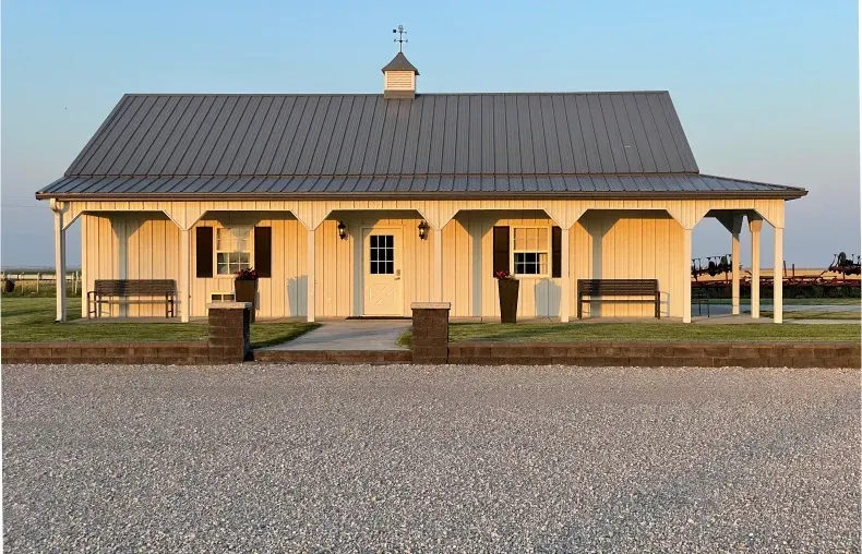 A white farmhouse-style building with a metal roof, front porch, and benches, set against a clear sky above gravel ground.