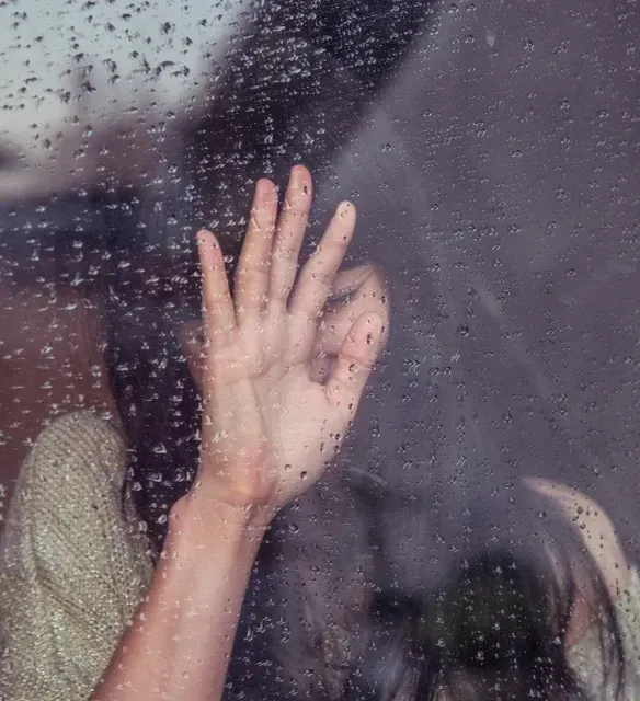 A person presses their hand against a rain-streaked window, their face softly blurred in the background.