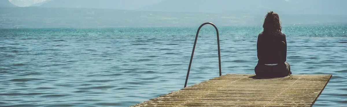 A person sits alone on the edge of a weathered wooden pier, looking out over a calm, vast body of water.