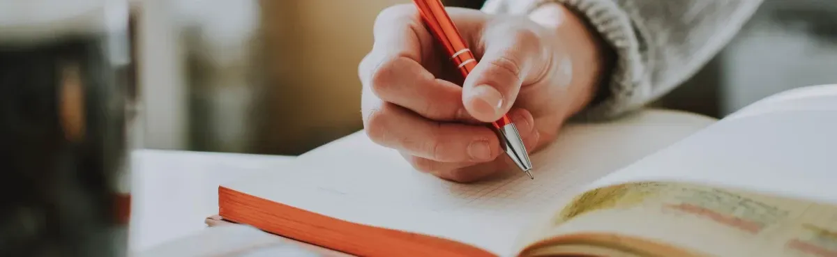 A close-up of a hand using an orange pen to write on an open notebook.