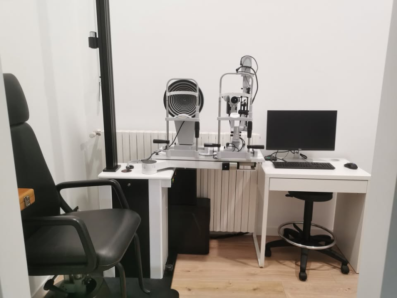 Sala visita oculistica con sedia, attrezzatura e computer. Pareti bianche e pavimento in legno.