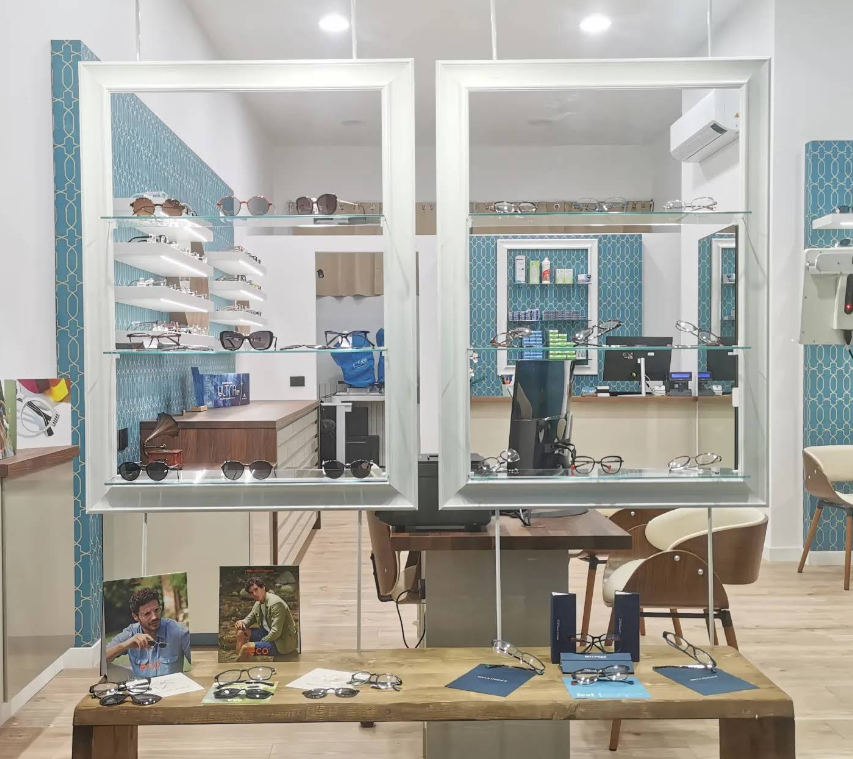 Interno di un negozio di occhiali con vetrine, occhiali e un tavolo in legno. Sfondo con piastrelle turchesi.