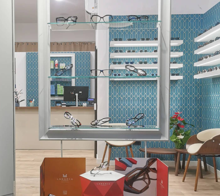 Interno di un negozio di ottica, espositori per occhiali, parete con motivi blu, pavimenti in legno, sedie e scatole per occhiali in primo piano.