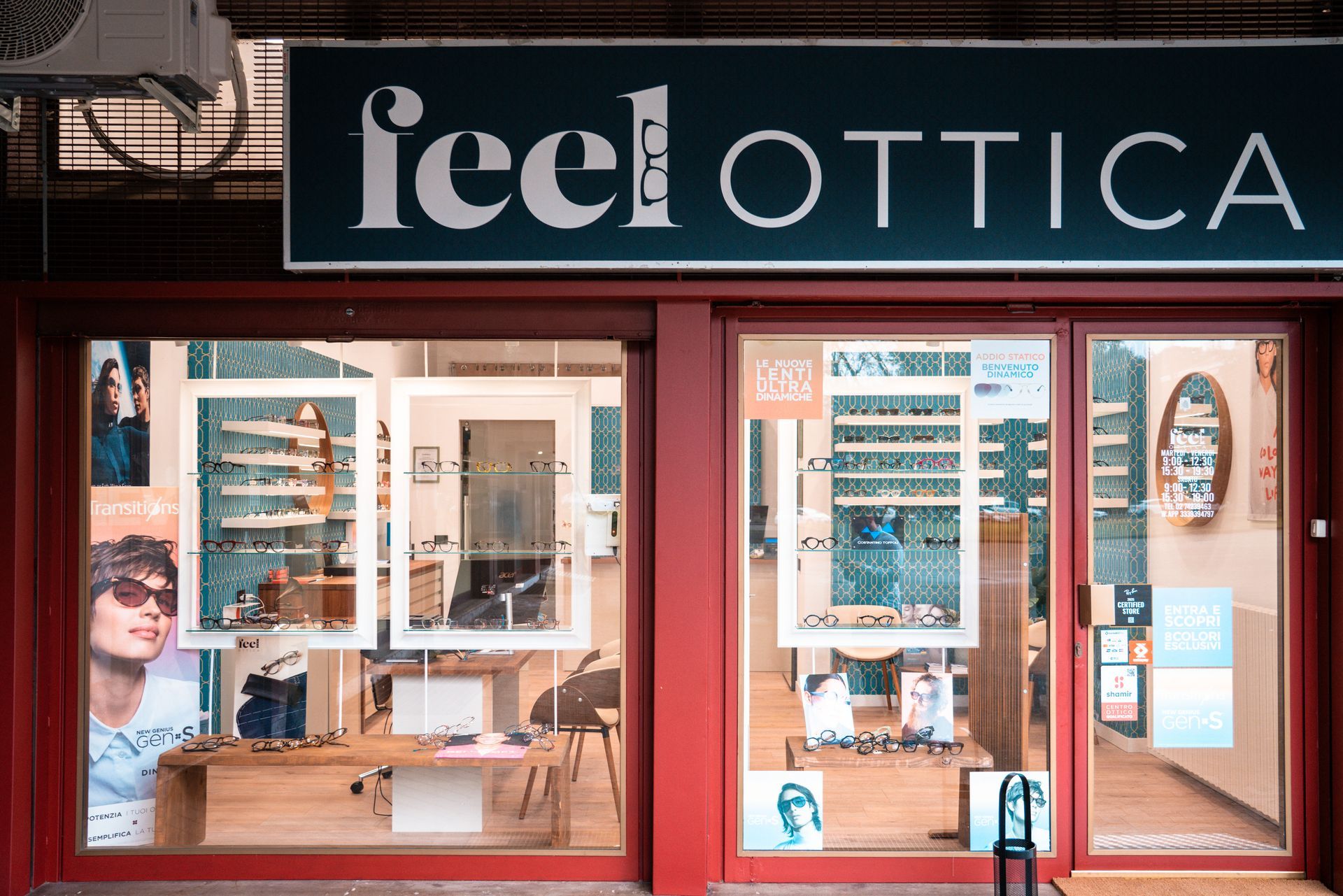 Interno di uno studio ottico con occhiali esposti su scaffali e sedie per i clienti. Carta da parati verde acqua.