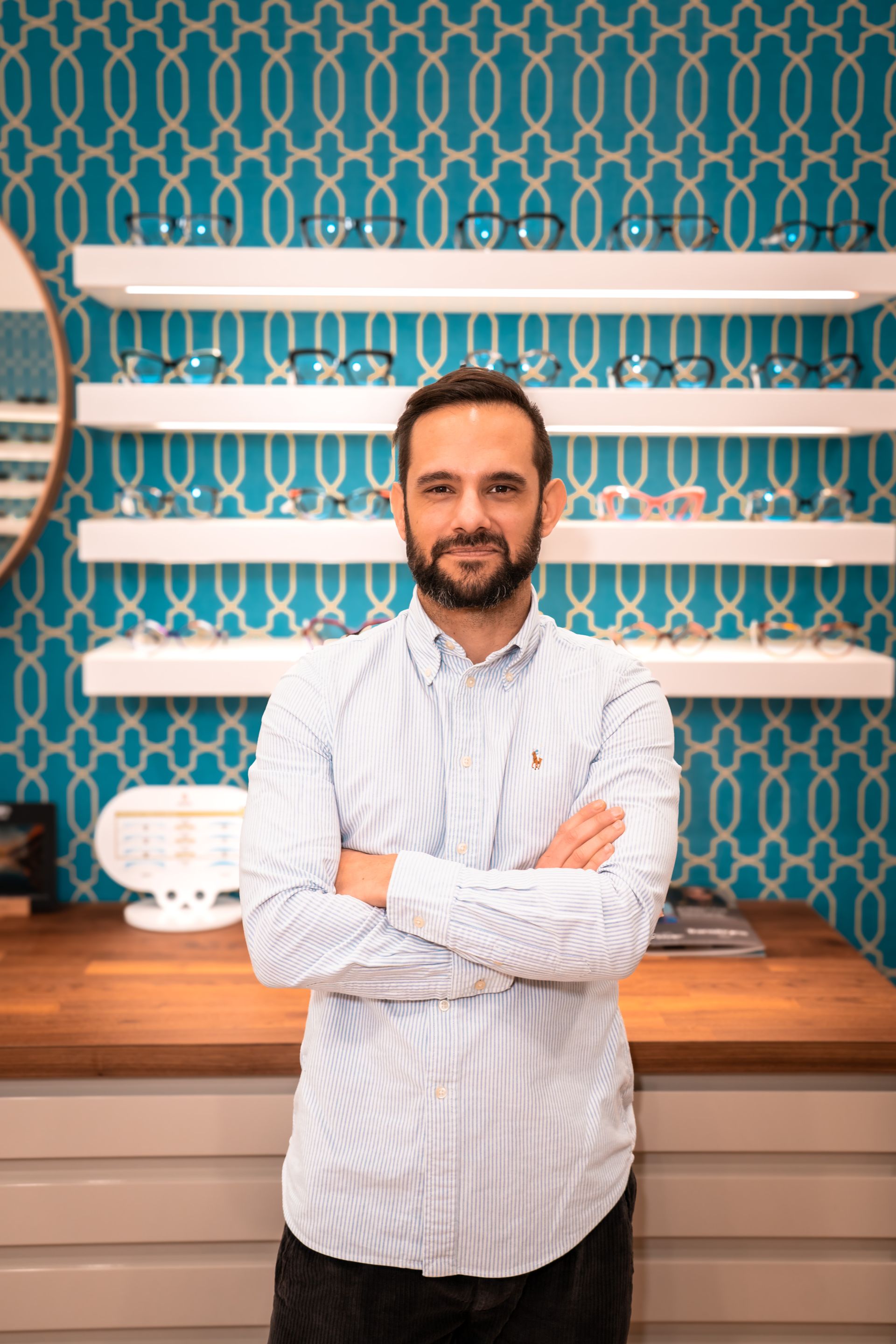 Interno di un negozio di ottica, espositori per occhiali, parete con motivi blu, pavimenti in legno, sedie e scatole per occhiali in primo piano.