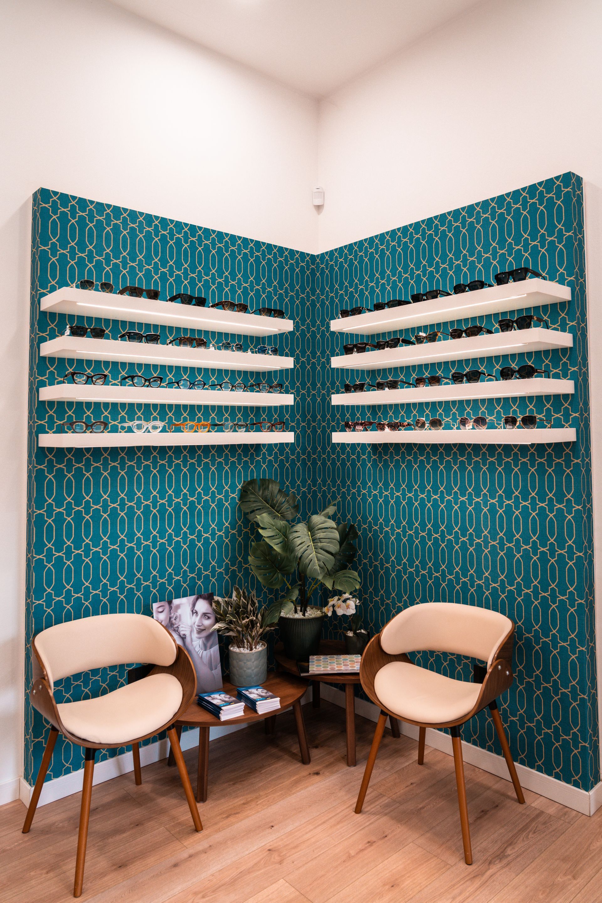 Interno di un negozio di ottica, espositori per occhiali, parete con motivi blu, pavimenti in legno, sedie e scatole per occhiali in primo piano.