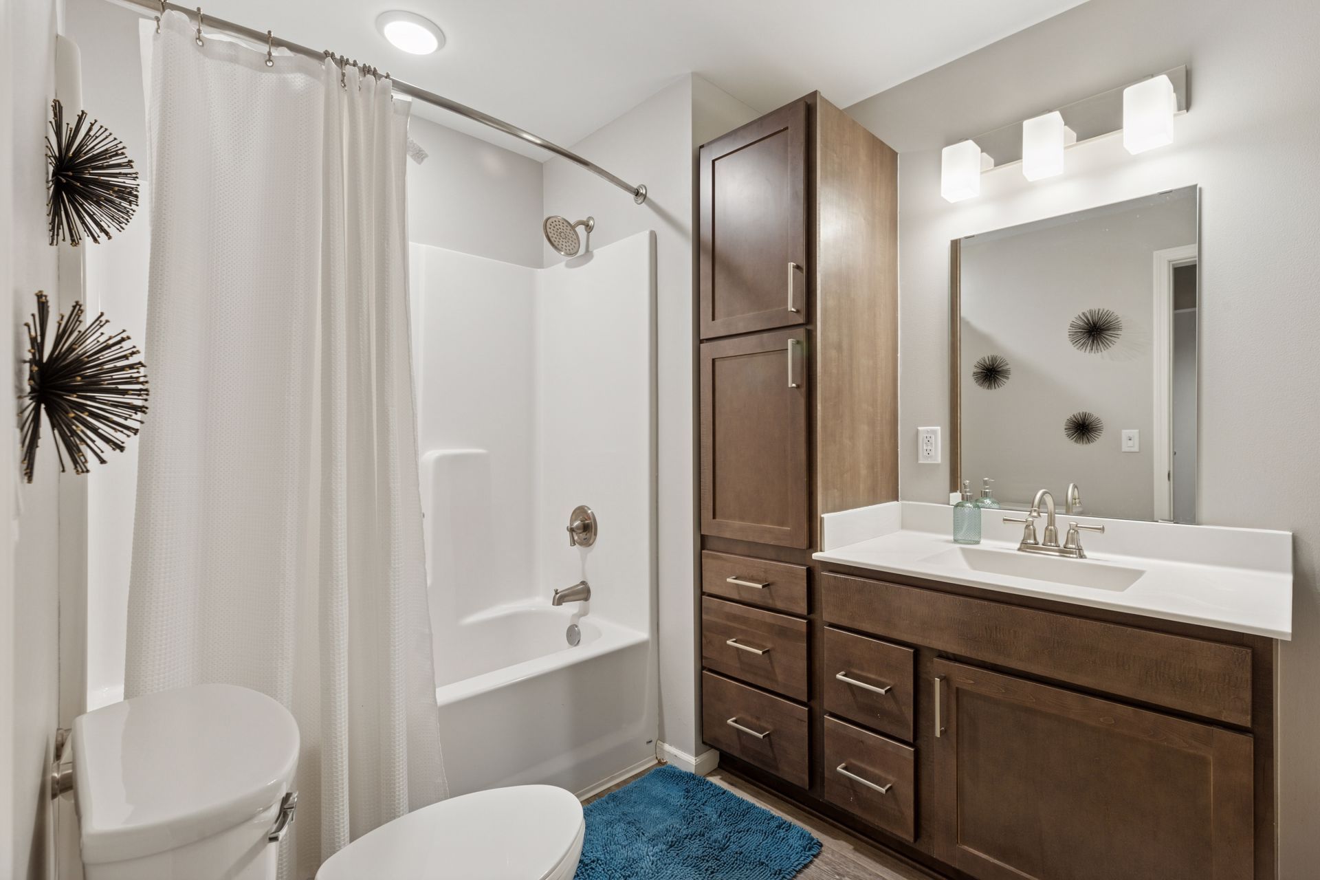 Bathroom with white shower, toilet, and vanity with brown cabinets. Blue bath mat, decorative wall art.