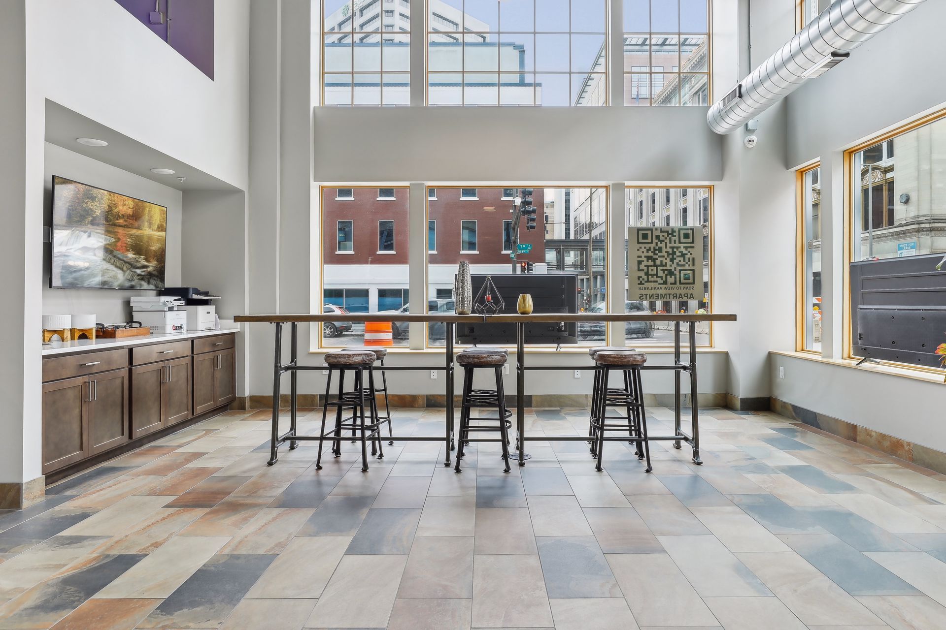Spacious room with a long table and stools in front of large windows overlooking a city street.