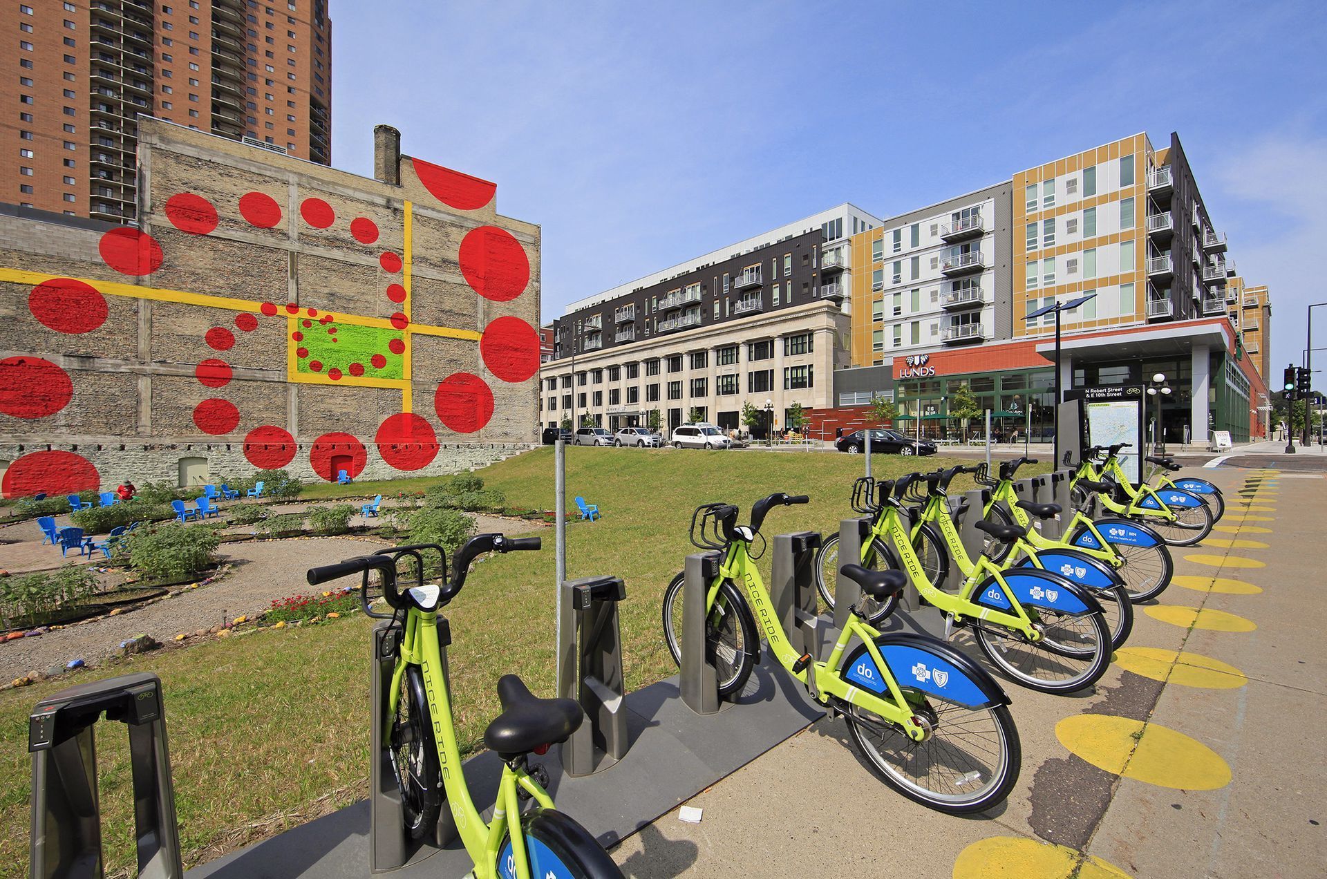 Green rental bikes docked on a sidewalk with a colorful mural and buildings in the background.