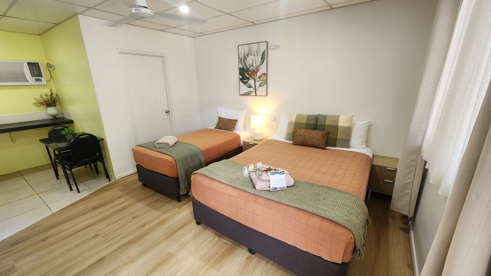 Two neatly made beds with peach and green bedding in a bright bedroom, separated by a wooden nightstand.