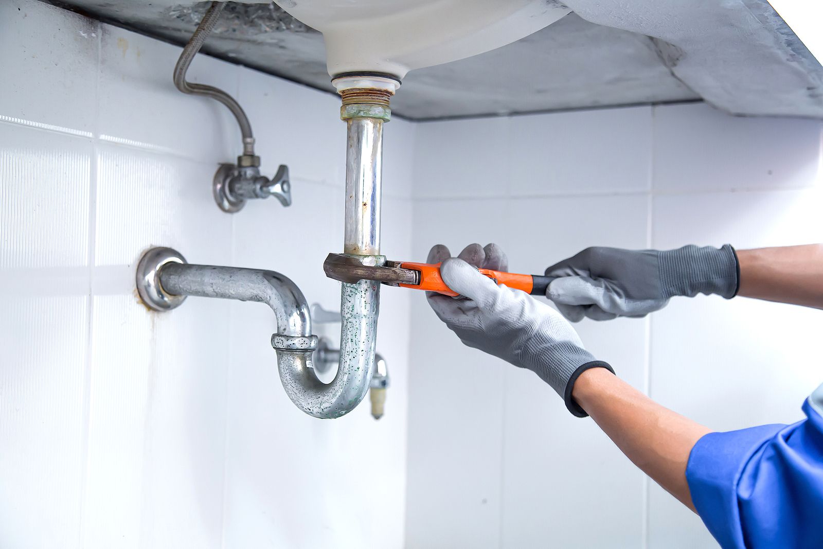 A person wearing gloves uses a wrench to work on plumbing under a sink in a tiled bathroom.
