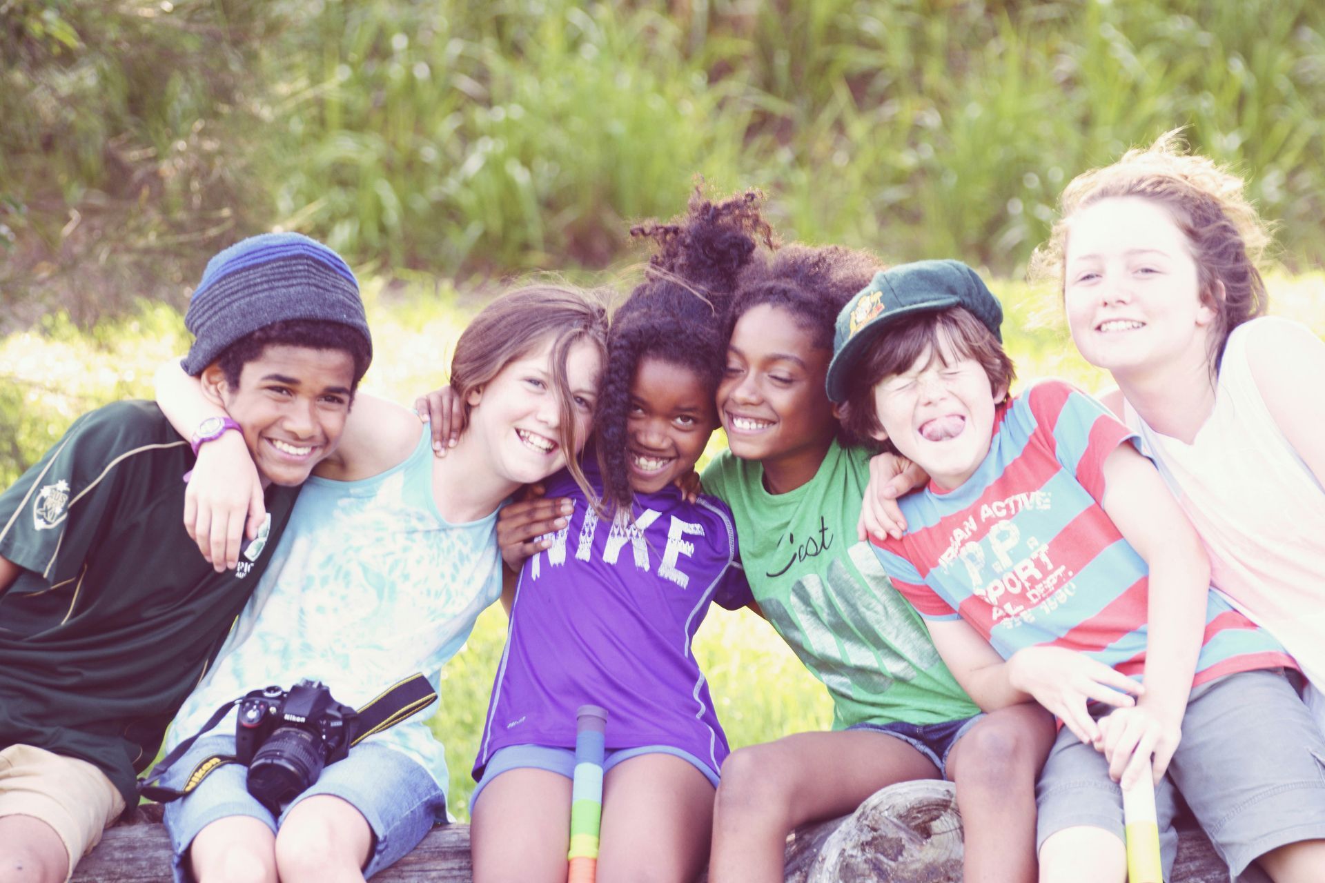 Six children sit closely together in a grassy field, smiling and hugging, with one child holding a camera.