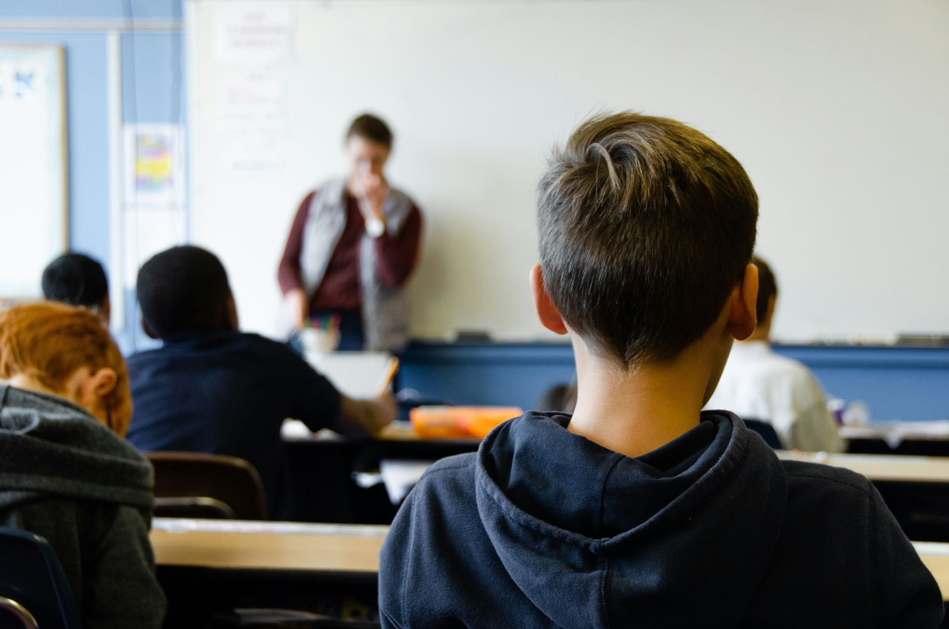 Students in classroom, facing away, teacher at whiteboard in background.