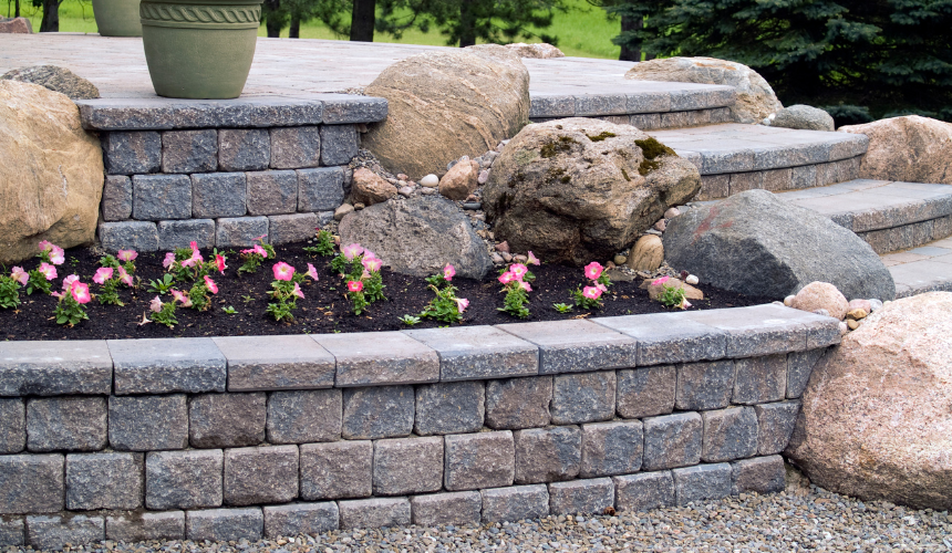 A stone patio with large boulders embedded into the stone patio and a raised garden bed featuring pink flowers.