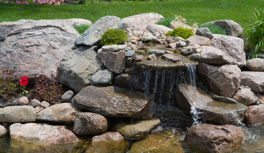 A serene single-layer waterfall cascades gently over natural rocks, flowing into a tranquil shallow pond—an oasis of calm and beauty in your garden.