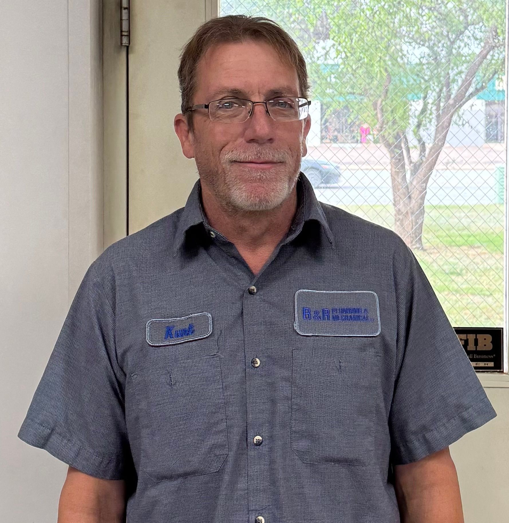 A man wearing glasses and a striped shirt stands in front of a window