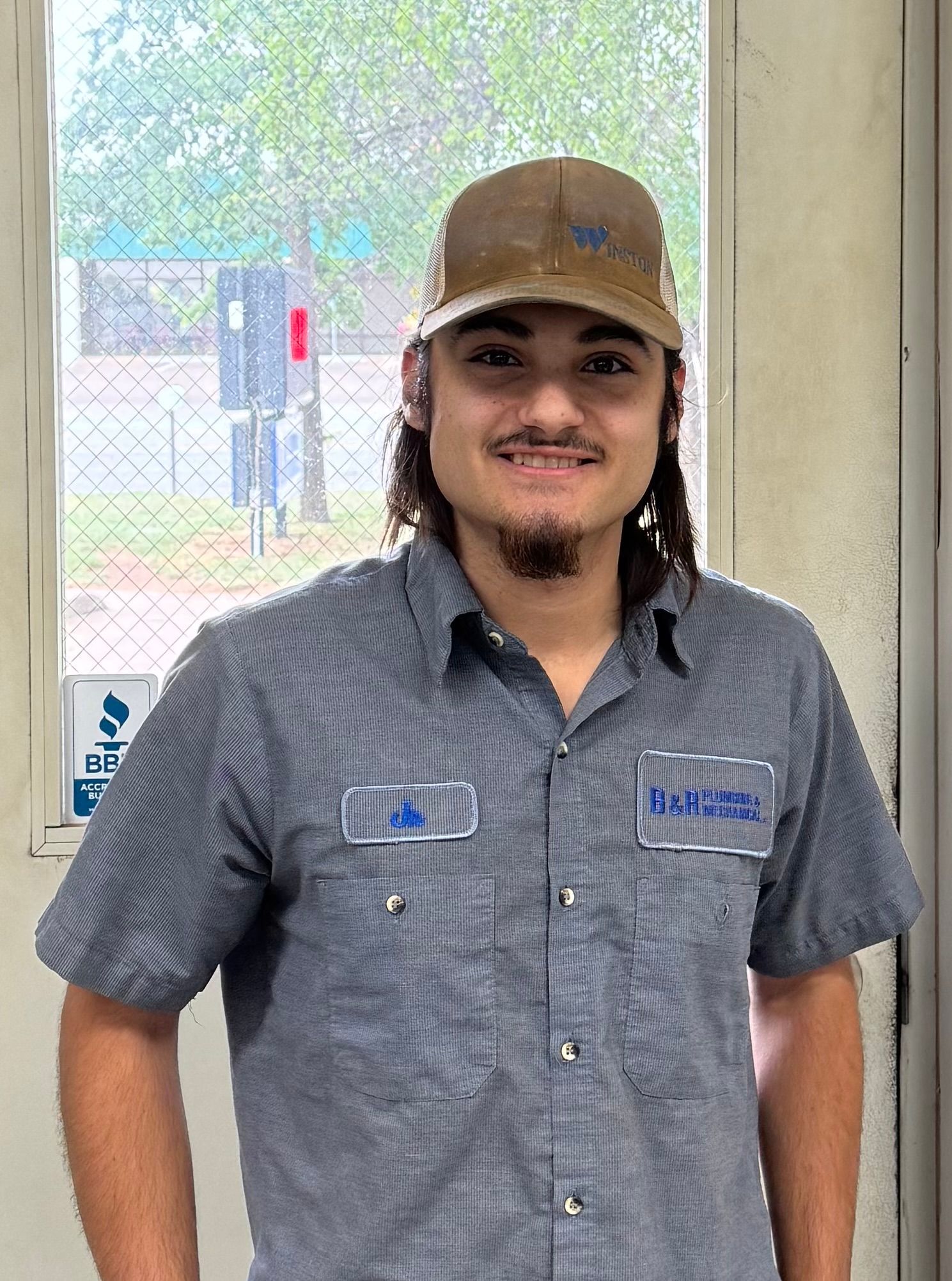 A man wearing a hat and a blue shirt is standing in front of a window.