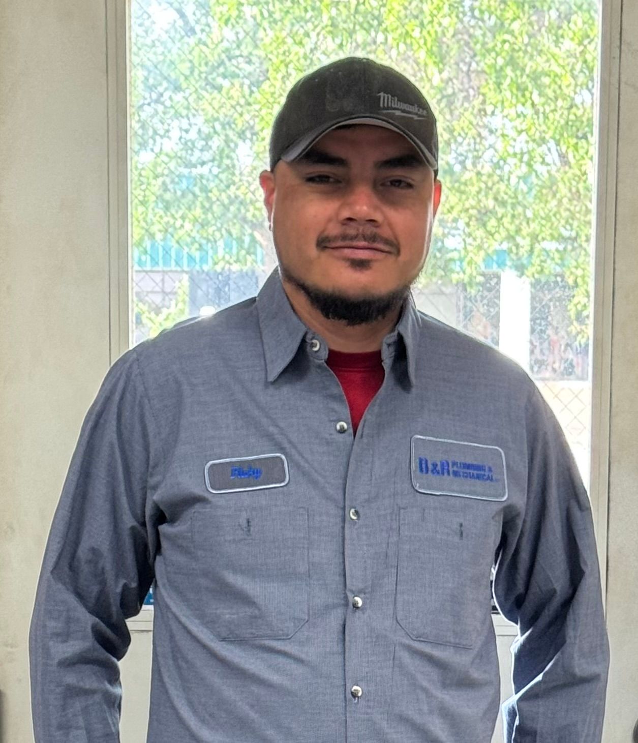 A man wearing a hat and a gray shirt is standing in front of a window.