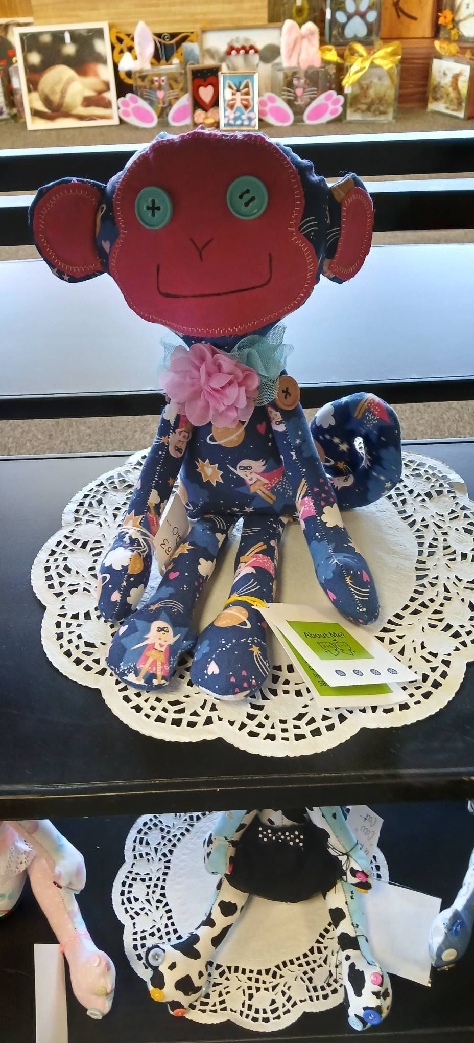 Red fabric doll with green button eyes and floral dress on a lace doily, shown on a shelf in a shop